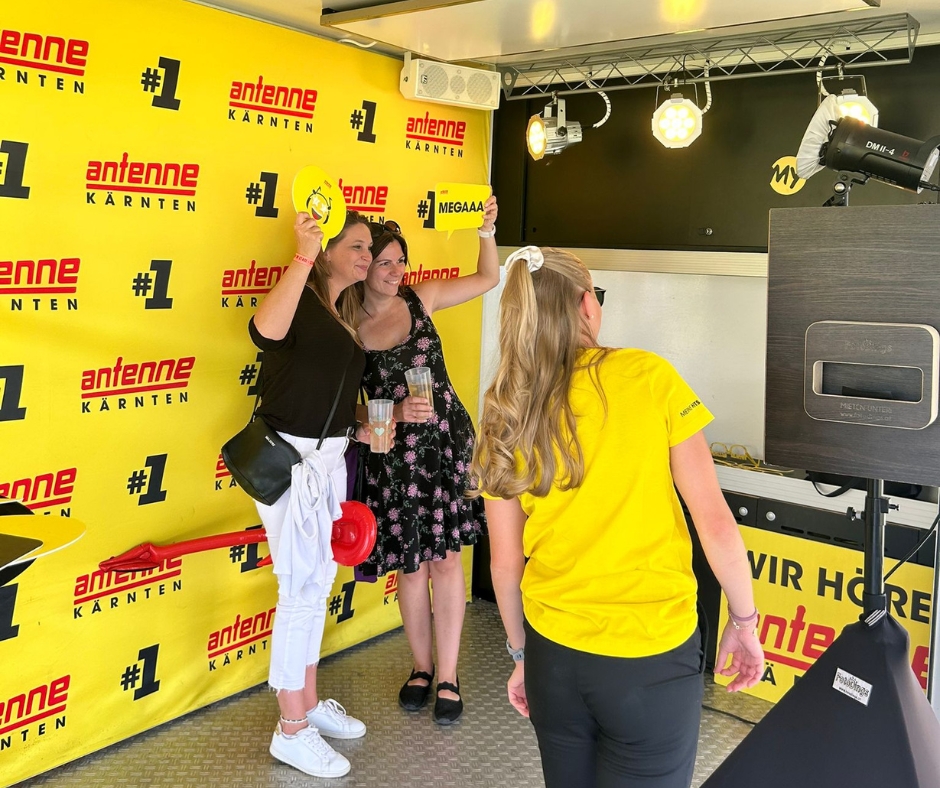 Zwei weibliche Gäste vor der Antenne Kärnten Werbewand bei einem Konzert mit einer UNIKAT Mitarbeiterin in Antenne Kärnten Gewand