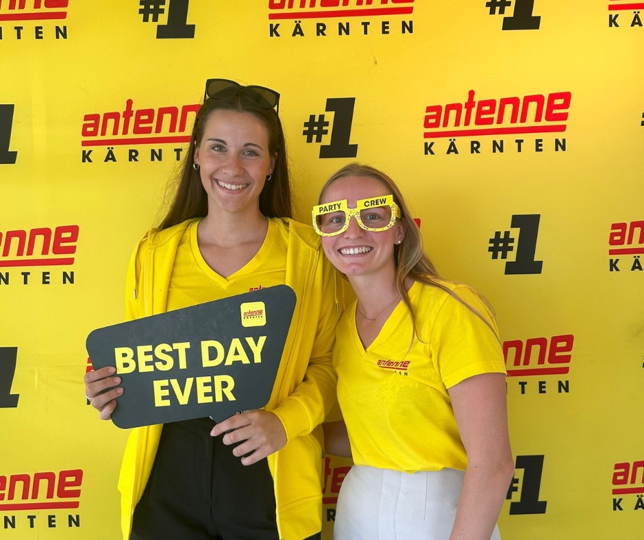 Zwei Frauen mit einem Schild "Best Day Ever" vor einer Antenne Kärnten Werbewand