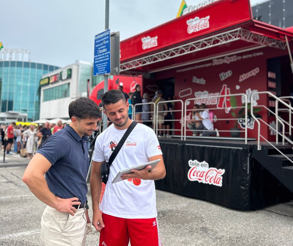 Zwei junge Männer, die auf ein iPad vor einer Coca Cola Bühne schauen.